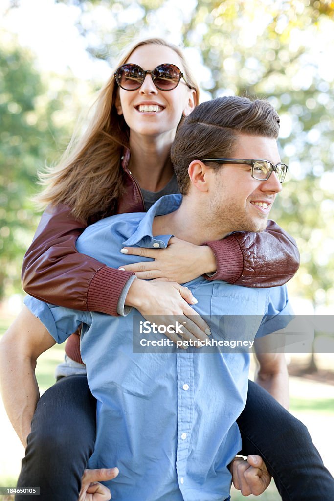 Young lady piggybacking on a man's back while both smile An attractive young couple in the park.  The guy gives the girl a piggy back ride. Eyeglasses Stock Photo