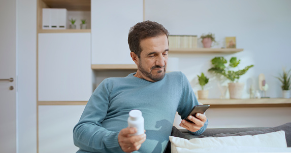 Senior man using smart phone while holding pill bottle at home.