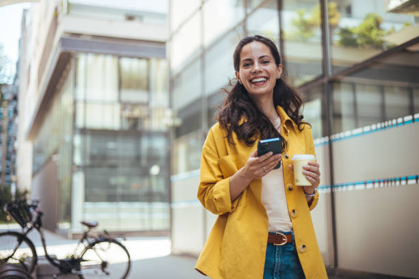 Woman texting and drinking coffee outdoors. Woman texting and drinking coffee outdoors. Young smiling brunette woman holding a smartphone and a cup of coffee looking at device screen against the background of an office building. liverpool one stock pictures, royalty-free photos & images