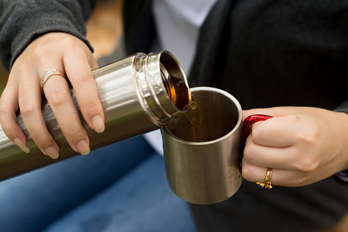 Image of an Asian Women sitting at the park while pouring hot tea using a stainless tumbler and  cup.
