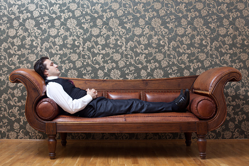 A young mesmerized man is lying down on brown leather coach in  psychiatrist office.The wall is wallpapered.The model and coach is seen in full length.He is in formal clothes and his hands are on his belly and his eyes are closed.The image was shot with a full frame DSLR camera.