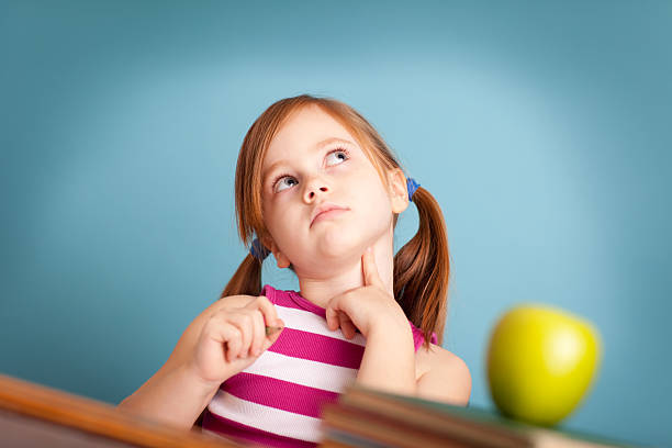 Color image of a serious young girl student sitting and thinking at a school desk on blue background.