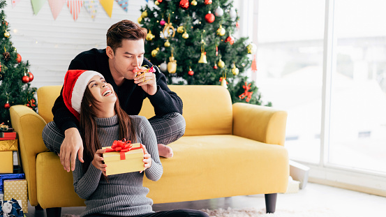 A young happy Asian man blowing a party whistle in the back at his girlfriend wearing a Santa Claus hat holding a received present at home with a Christmas tree in the background.