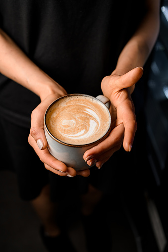 Woman holding in hands cup with tasty steaming cappuccino decorated with foamy art, top view. Hot served coffee in woman's hands