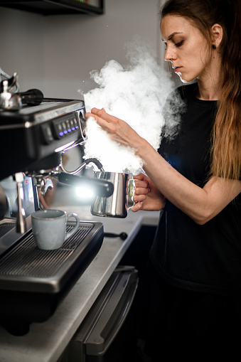 Side view of young woman barista frothing milk for latte in coffee machine. Barista working with professional coffee machine at coffee shop