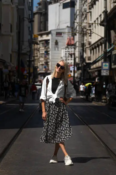 Photo of Beautiful blonde young woman in sunglasses poses on one of the main streets of Istanbul on a sunny day. Stylish female tourist with a backpack in Turkey. Vertical photo