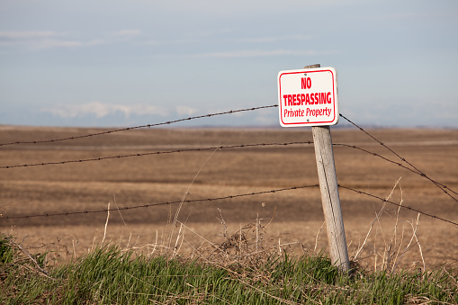 A no trespassing sign on the prairie. This image of a no trespassing private property sign with a barbed wire fence is taken near Calgary, Alberta on a rural property. Trespassing can be a charge with severe consequences as landowners often take the law into their own hands! Respecting the rights of others, especially their personal property, is a requirement of every citizen. This image was taken in spring before this field was planted. 