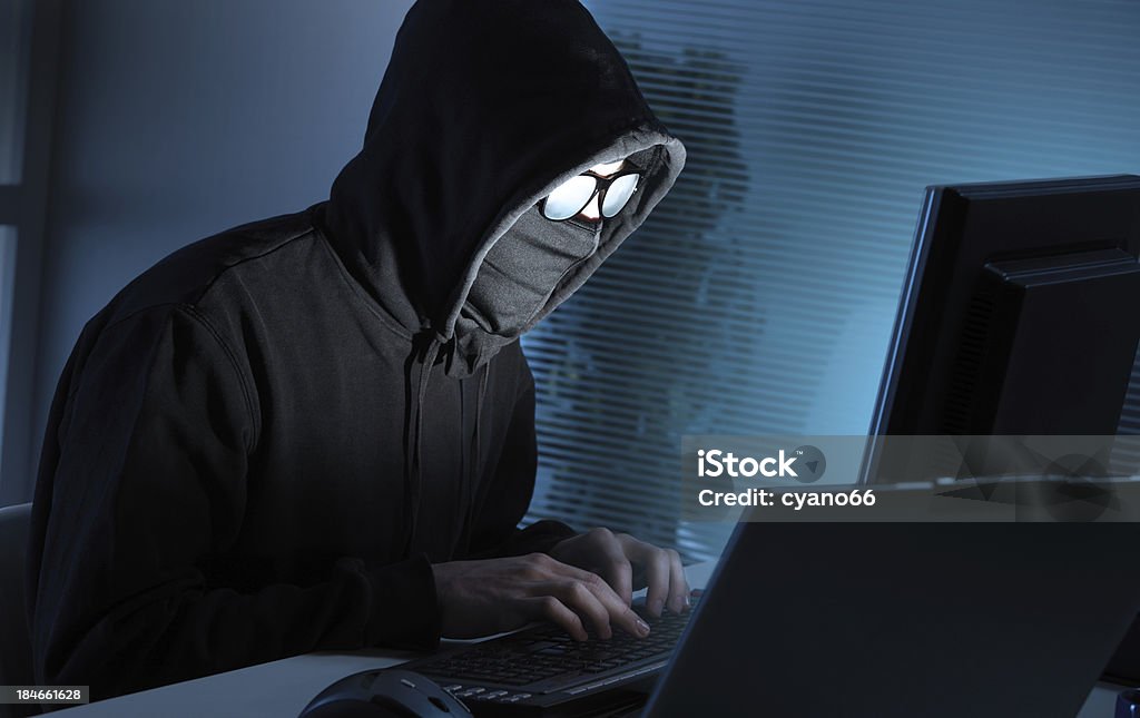 Hacker stealing data from computer Young hacker in the act of stealing data from a computer.  The young man is wearing a hoodie that covers his head, glasses and a mask over his face.  He is sitting at a desk in front of a desktop computer.   Next to the computer is a laptop and blinds in the background. Computer Hacker Stock Photo