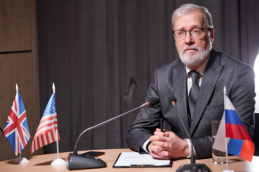 senior businessman sitting with microphone in modern boardroom during meeting with business partners, handsome male in eyeglasses confidently sitting, wearing formal suit