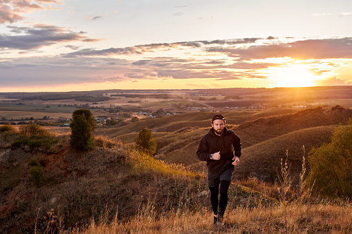 caucasian man jogging along the field, wearing sportive clothes, practice running outdoors in the morning at sunrise, in nature. young male engaged in sport, motivated. strong and healthy person