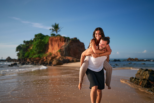 Couple enjoys piggyback on sandy beach. Man carries girlfriend on back, smiles by sea near cliff. Holidaymakers share joy, love on tropical coast. Pair experience romantic getaway, ocean adventure.