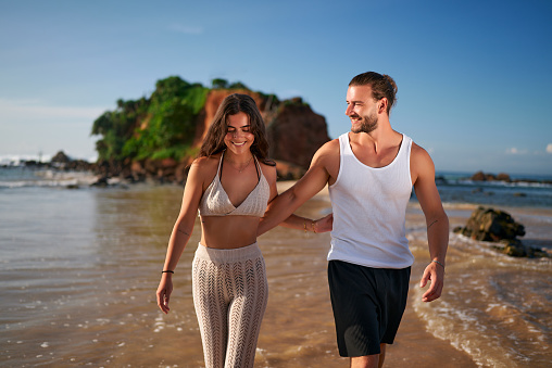 Happy couple walks on beach, holding hands, smiling, barefoot near sea. Man, woman in summer outfits enjoy sunshine, coastal lifestyle. Remote work concept, travel, leisure, relationship goals.