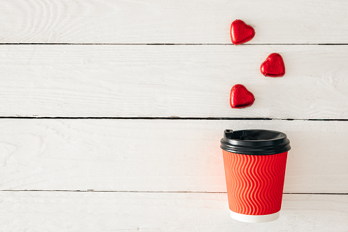 Red paper cup and heart-shaped candies on a white wooden background. View from above. Space for text. Valentine's Day concept.