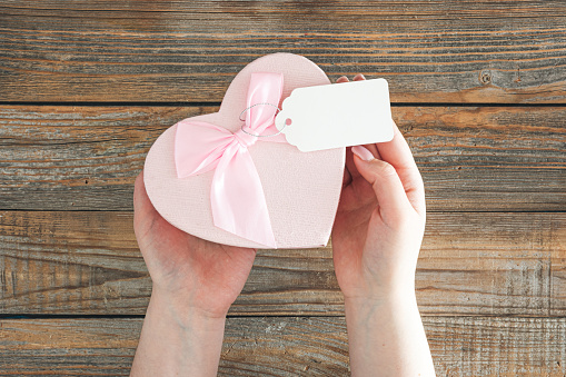 Pink heart shaped gift box with blank gift tag in female hands on wooden background. Valentine's Day concept.