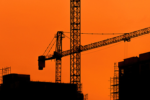 Silhouette of construction crane, scaffolding and buildings in sunset against orange sky. Copy space included.