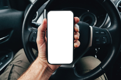 Driver app mockup with blank smartphone screen over steering wheel, man holding mobile phone device in car, selective focus