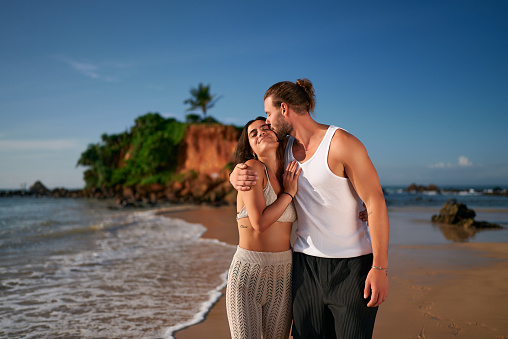 Affectionate couple embrace on tropical beach at sunset. Childfree travelers enjoy serene sea, sandy shore. Young man, woman experience romantic getaway. Love, travel, leisure without kids depicted.