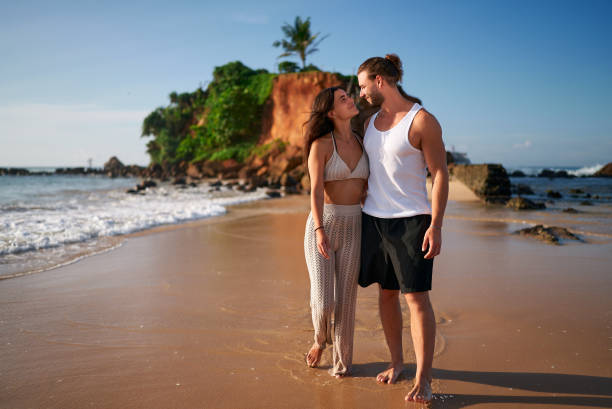 istock Romantic pair strolls on sandy beach by sea at sunset. Affectionate man, woman walk barefoot, ocean waves wash feet. Couple enjoys seaside, embraces. Travelers adore coastal landscape, tranquility. 1846764893