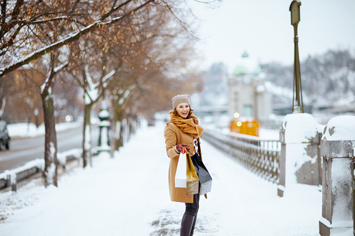 smiling modern woman in brown hat and scarf in camel coat with gloves and shopping bags outside in the city in winter.