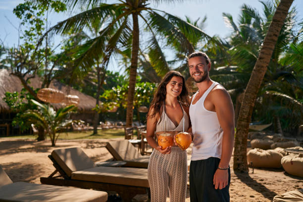 Smiling couple enjoys honeymoon in tropical paradise, sharing fresh coconut water. Fit man and woman in summer attire stand amidst palm trees, celebrating romantic getaway with exotic drinks. Smiling couple enjoys honeymoon in tropical paradise, sharing fresh coconut water. Fit man and woman in summer attire stand amidst palm trees, celebrating romantic getaway with exotic drinks. summer stock pictures, royalty-free photos & images