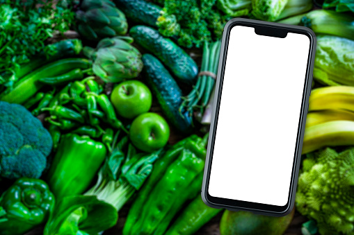 Mobile phone with blank screen on green fruits and vegetables background. Copy space