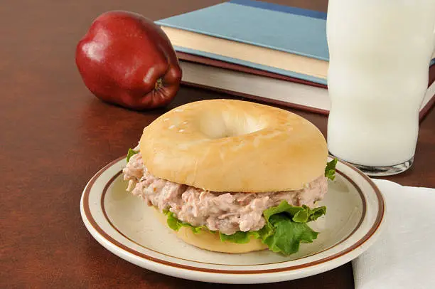 A tuna sandwich on a bagel with an apple, glass of milk and school books