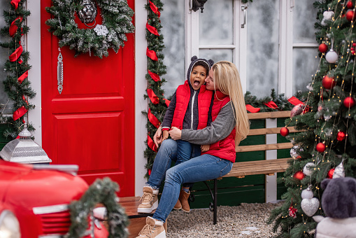 Caucasian mother with African American son having fun in backyard, on bench near red door with wreath. Young blonde woman kisses little boy, tickles him, the diversity family laughs at Christmas tree