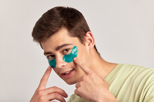Young handsome smiling guy with brown hair applying under eye hydrogel patches on his face. Caucasian millennial man practices skin care routine to keep healthy and youthful looking. Studio portrait.