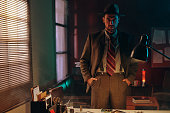 Serious man in retro suit and hat standing in dark office and looking at you
