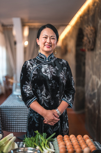 Portrait of a beautiful Chinese mature female chef dressed in traditional black Chinese Cheongsam smiling in front of the camera in the restaurant with fresh ingredients on the checked patterned table.
