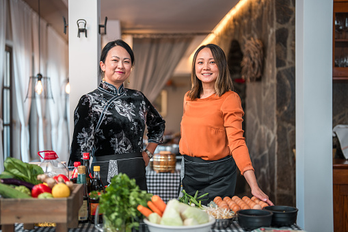 Portraits of beautiful Chinese mature female chefs dressed in traditional black Chinese Cheongsam and modern orange top posing in front of the camera in the restaurant with fresh ingredients on the checked patterned table.