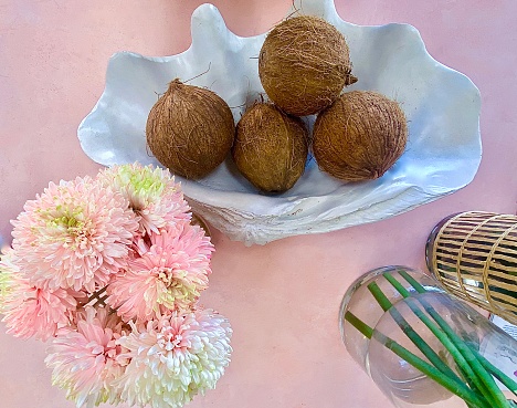 Horizontal flat lay of pastel pink kitchen table with large shell holding whole coconuts next to dahlia flowers and vases in country Australia