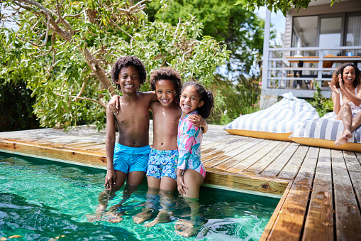 Portrait of a two little brothers and their young sister smiling while standing together in their swimming pool at home in summer with their mom watching