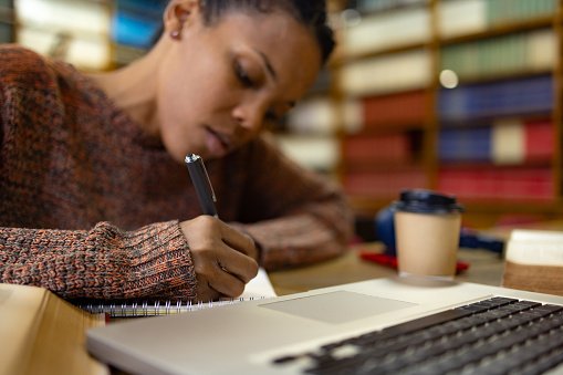 Close up of African American college student writing in a notepad while studying in library.