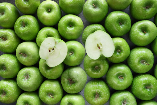 Stock photo showing close-up view of a halved apple on top of a group of green Granny Smith apples with shiny, speckled skin.