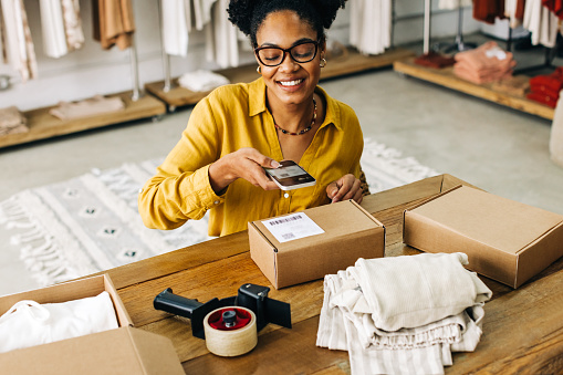 Fulfilling clothing orders for her online store, this female dropshipper scans a QR code with her mobile phone, showcasing her entrepreneurship skills and dedication to customer satisfaction.