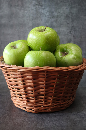 Stock photo showing close-up view of a group of green Granny Smith apples with shiny, speckled skin heaped high in a wicker basket against a  mottled dark grey background.