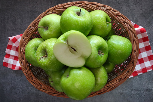 Stock photo showing close-up, elevated view of a group of green Granny Smith apples with shiny, speckled skin heaped high in a wicker basket against a mottled dark grey background.