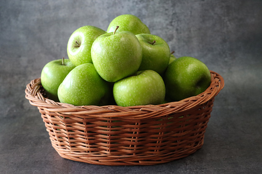 Stock photo showing close-up view of a group of green Granny Smith apples with shiny, speckled skin heaped high in a wicker basket against a  mottled dark grey background.