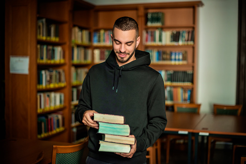 Young man standing in the university library and holding books in his hand.