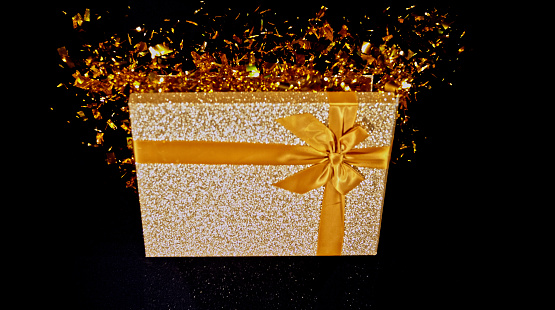 Close-up of gold coloured gift box exploding with confetti against black background.
