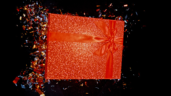 Close-up of red gift box exploding with confetti against black background.