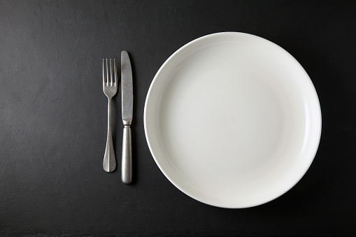 Empty white dinner plate and cutlery on a black stone table. Top view with copy space.