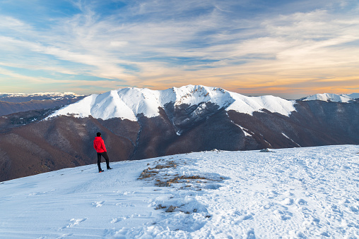 One person with red jacket standing on top of a hill admiring snow covered mountains at sunset, Abruzzo, Apennines, Italy, Europe