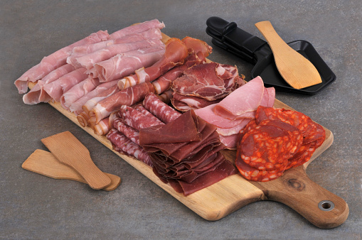 Assorted cold meat slices on cutting board with kitchen utensils on gray background