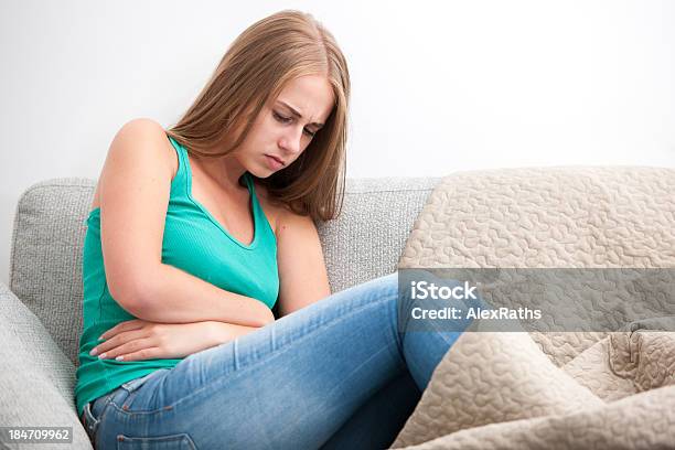 Young Girl Sitting On A Sofa Holding Her Stomach In Pain Stock Photo - Download Image Now