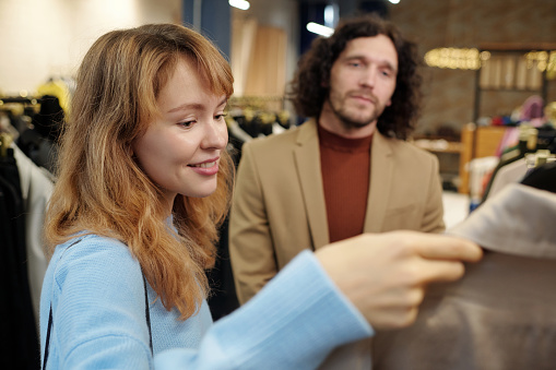 Young blond woman in blue pullover looking at coat on hanger while choosing new attire from seasonal collection with her husband