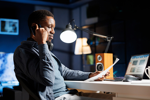 Working from home to expand enterprise on global stage, african american man calls creditors to acquire more money for capital expenditure. At night, staff member talks on mobile device.