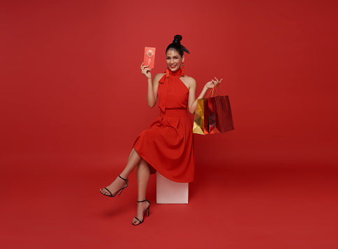 Happy asian woman in red dress holding shopping bags and gift voucher for Chinese new year shopping sitting on chair isolated on red background. Chinese text means great luck great happy.