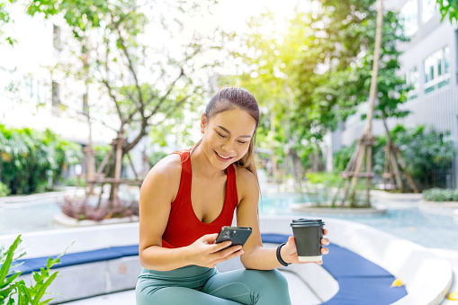 Asian Woman Sitting in the Park, Using Smartphone and Holding a Cup of Coffee After Exercise. Digital banking anytime anywhere. Business on the go.